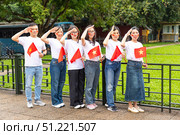 Hanoi, Vietnam - 29.04.2025: A group of six young people stand on a street, holding Vietnamese flags in one hand and saluting near their heads with other hand. National holiday and patriotism concept. Редакционное фото, фотограф Ирина Михайличенко / Фотобанк Лори