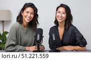 Female podcasters smiling, recording radio show in professional studio, wearing headphones and sharing enthusiastic conversation near condenser microphones. Стоковое AI-изображение, AI-художник Юрий Поздников / Фотобанк Лори