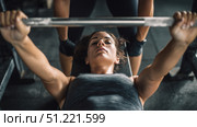 Young woman bodybuilder lifting a barbell with focused determination, guided by her personal trainer in a dimly lit gym, showcasing strength and fitness. Стоковое AI-изображение, AI-художник Юрий Поздников / Фотобанк Лори