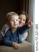 Cheerful toddler pointing skyward as caring mother observes nearby, capturing tender family interaction within soft, inviting domestic setting. Стоковое AI-изображение, AI-художник Юрий Поздников / Фотобанк Лори