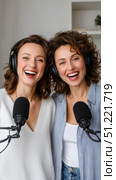 Female radio hosts giggling, wearing headphones while recording podcast together using professional microphones in cozy home studio setting. Стоковое AI-изображение, AI-художник Юрий Поздников / Фотобанк Лори