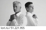 Sophisticated father son duo adjusting crisp white shirts, posing back to back against minimalist white backdrop in monochrome fashion shoot. Стоковое AI-видео, аниматор Юрий Поздников / Фотобанк Лори