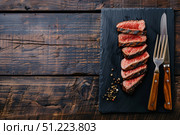 Крупный план приготовленного мясного стейка, поданного ломтиками на прямоугольной черной доске с перцем и солью, на фоне старого дерева. Стоковое AI-изображение, AI-художник Кирилл Сиротюк / Фотобанк Лори