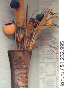 Interior composition of dried flowers in a decorative vase. The elongated vase is made in brown tones with a relief leaf pattern, giving it a natural and handmade character. The bouquet contains various dried plants: ears of corn, twigs, seed pods, woody elements and decorative figured inserts. The color scheme of the bouquet is maintained in natural beige, brown and golden shades with accents of dark blue dried flowers. The composition is located against the background of a textured wall with a pattern, which emphasizes its decorative and ethnic sound. Such a design is used to decorate interiors in the style of eco, boho, rustic or vintage. Стоковое фото, фотограф Анна Кривицкая / Фотобанк Лори