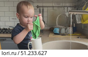 A toddler is happily washing a cup in the kitchen while enjoying the running water playfully. Стоковое видео, видеограф Olesia Kondrateva / Фотобанк Лори