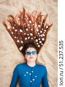Stylish summer portrait of young girl lying on sand with seashells in her long hair, wearing heart-shaped sunglasses. Concept of creative holiday vibes, beach lifestyle, and youth fashion. Стоковое фото, фотограф Енваев Андрей / Фотобанк Лори