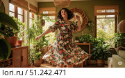 Happy lifestyle concept with beautiful woman dancing in cozy home interior, carefree female in floral dress enjoying weekend in living room with many plants. Стоковое AI-изображение, AI-художник Павел Муравьев / Фотобанк Лори