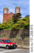 Nagasaki, Japan - May 12, 2025. Red Taxi on the road and Urakami Church on Back. Стоковое фото, фотограф Константин Никитеев / Фотобанк Лори