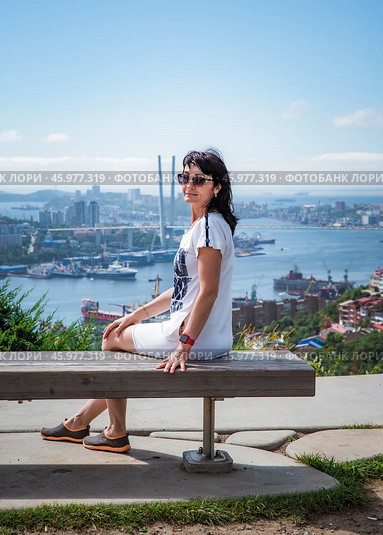 A 50-year-old Russian woman in a white dress sits on a bench in Nagorny Park, Vladivostok, looking at the camera with Golden Horn Bay in the background. Стоковое фото, фотограф Татьяна Терехина / Фотобанк Лори