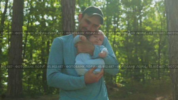 A bearded, joyful father in a baseball cap and blue jersey holds an infant dressed in a blue onesie. The father cradles his head against his son's head and kisses him tenderly. Family in nature. Стоковое видео, видеограф Воловодов Владимир / Фотобанк Лори