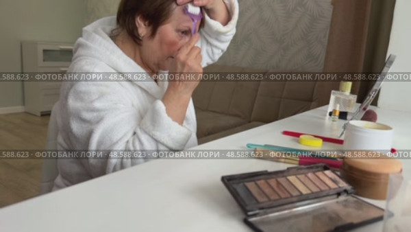 A beautiful senior woman is sitting at home and doing her make-up morning routine. Senior woman putting blush on her face. Стоковое видео, видеограф Ирина Старикова / Фотобанк Лори