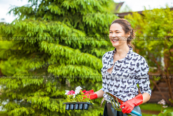A beautiful woman laughs cheerfully and holds garden tools and flowers in her hands. Стоковое фото, фотограф Astroid / Фотобанк Лори