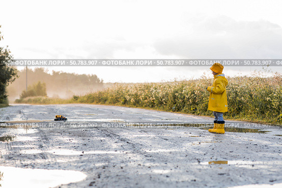 A boy in yellow rubber boots and a raincoat runs through puddles, having fun and playing with a remote-controlled car after it rains. Summer and autumn holidays. A happy childhood. Стоковое фото, фотограф Photojuli / Фотобанк Лори