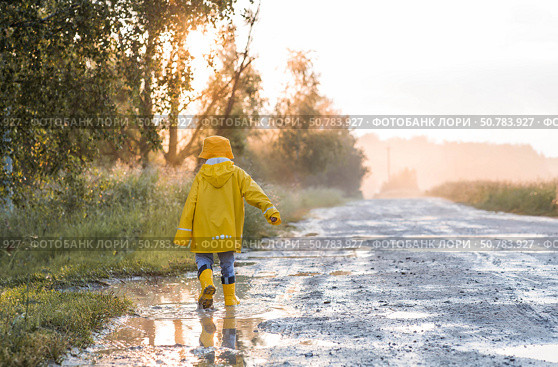 A boy in yellow rubber boots and a raincoat runs through puddles, having fun and playing after it rains. Summer and autumn holidays. A happy childhood. Стоковое фото, фотограф Photojuli / Фотобанк Лори