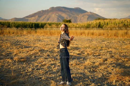 A carefree woman stands joyfully in a vast golden field, her arms wide open as she embraces the stunning beauty of the majestic mountains, which are illuminated by the warm, golden rays of the sunset. Joyful Woman in Nature Embracing the Essence of Freedom in a Beautiful Scenic Landscape. Стоковое фото, агентство Ingram Publishing / Фотобанк Лори