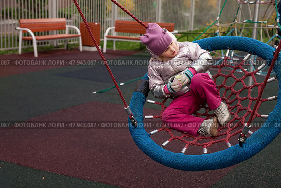 A cheerful five-year-old girl enjoys swinging on a colorful net swing at an autumn playground. Surrounded by bright foliage, she expresses curiosity and joy while engaging with her environment. Стоковое фото, фотограф Aleksandr Golubev / Фотобанк Лори