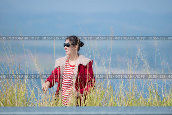 A cheerful young woman in a red jacket stands on top of a green hill, arms spread. Стоковое фото, агентство Ingram Publishing / Фотобанк Лори