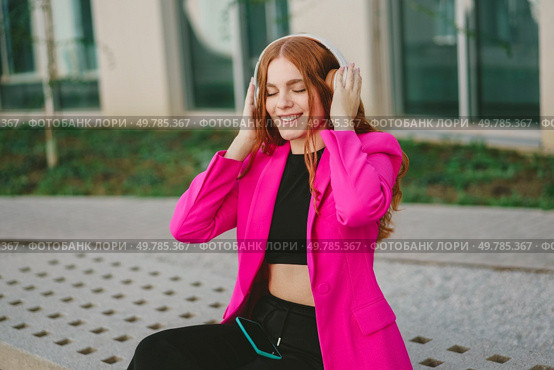 A cheerful young woman sporting long hair and a vibrant pink blazer, paired with trendy headphones, joyfully enjoys her favorite music outdoors, radiating positive energy, happiness, and unique style. A young woman is enjoying her favorite music while dressed in a stylish pink blazer. Стоковое фото, агентство Ingram Publishing / Фотобанк Лори