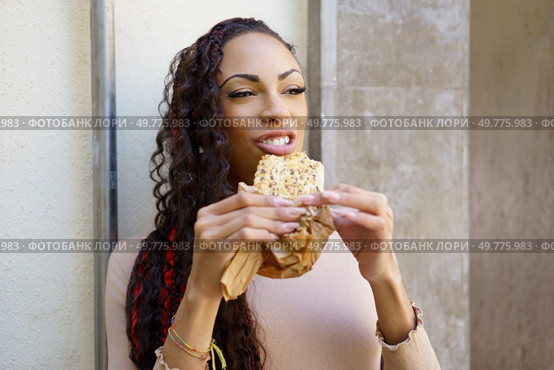 A cheerful young woman with curly hair beams with delight as she takes a satisfying bite of a delicious snack that is wrapped in paper, cherishing her wonderful moment spent outdoors in nature. A woman is enjoying a truly delicious and tasty snack while being outdoors in the fresh air. Стоковое фото, агентство Ingram Publishing / Фотобанк Лори