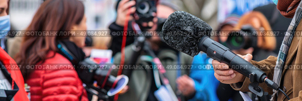 A close-up image of a journalist holding a microphone while interviewing someone at an outdoor event, surrounded by other journalists and members of the public. Стоковое AI-изображение, AI-художник Данила Большаков / Фотобанк Лори
