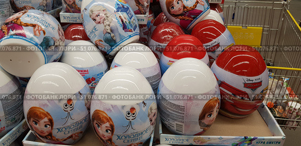 A close-up of a shop window or product display. In the foreground are large plastic eggs with images of Disney and Pixar characters. The blue and white eggs are Frozen-themed with images of Anna, Elsa, and Olaf. The red and white eggs are Cars-themed with images of Lightning McQueen. These eggs usually contain sweets and toys, which attracts children. The products are neatly packed into boxes, some of which are in a trolley. The background is the retail space of the shop. (2018 год). Редакционное фото, фотограф Анна Кривицкая / Фотобанк Лори
