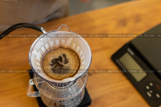 A coffee dripper sits on a scale, with freshly brewed coffee dripping into a glass carafe. The scene features a wooden table and a digital scale. Стоковое фото, фотограф PAv / Фотобанк Лори