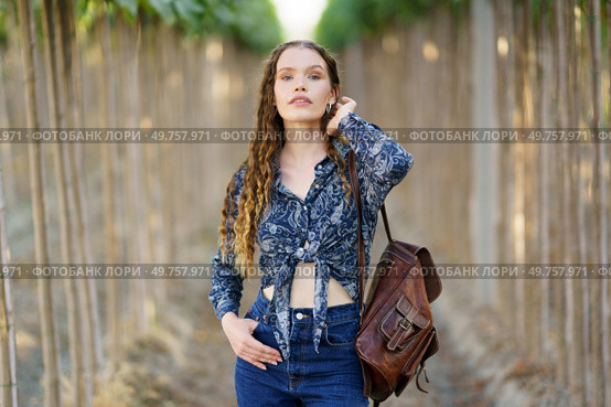 A confident young woman in a stylish denim outfit holds a chic backpack, surrounded by serene nature. Her bright smile and relaxed demeanor reflect a carefree summer lifestyle, embodying adventure. A Confident Young Woman Dressed in Trendy Denim with a Stylish Backpack in a Serene Nature Setting. Стоковое фото, агентство Ingram Publishing / Фотобанк Лори
