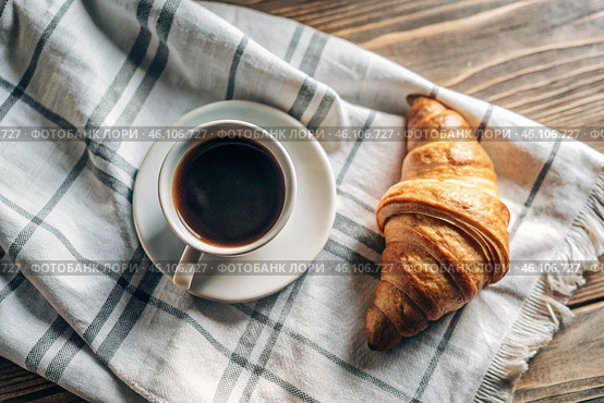 A delicious freshly baked croissant, a white cup of coffee and a cotton napkin on a wooden background. French breakfast. Delicious pastries in close-up. Стоковое AI-изображение, AI-художник Photojuli / Фотобанк Лори