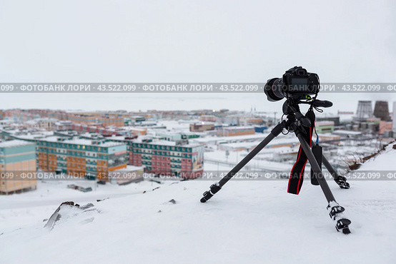 A digital camera on a tripod photographs a snow-covered northern city in the Arctic. Tourism and winter photography in the Far North. Cold snowy weather. Travel photography equipment. Photo hobby. Стоковое фото, фотограф Андрей Степанов / Фотобанк Лори