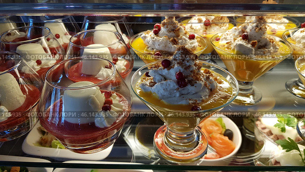 A display case with exquisite desserts in glass goblets. The focus is on two types of desserts: one with panna cotta, decorated with red berry sauce and lingonberries, the other with a delicate mango or citrus mousse, generously decorated with whipped cream, nut crumbles and berries. The desserts look fresh, airy and appetizing. At the bottom of the frame, elements of the appetizers are visible, including salmon and greens, which creates a contrast effect between sweet and salty. Стоковое фото, фотограф Анна Кривицкая / Фотобанк Лори