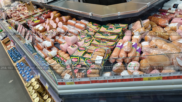 A display refrigerator in a supermarket filled with various types of sausages. The display case includes cooked sausages, frankfurters, wieners, smoked and semi-smoked sausages, as well as sliced ​​sausages in vacuum packaging. The products are neatly laid out: in the foreground, sausages and wieners in film are visible, behind them are loaves of cooked sausage and packages with slices. The packages have bright green, yellow and red labels. Under the glass are price tags with current prices for the products. At the bottom of the photo, glass jars of mustard and packages of bottled drinks are visible. The atmosphere of a typical supermarket with a large selection of meat products. Редакционное фото, фотограф Анна Кривицкая / Фотобанк Лори
