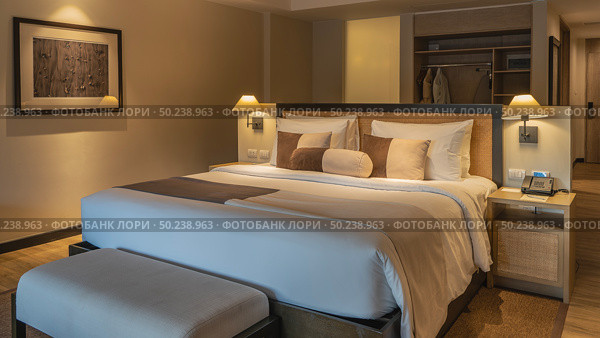 A double bed with white sheets and a beige bedspread. Pillows at the headboard. The couch. The phone is on the bedside table. The lamps are lit. The interior of a bedroom in a hotel. The suite. Стоковое фото, фотограф Вера Ивановна Кубарева / Фотобанк Лори