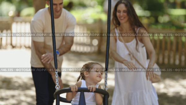 A family spending time in the park - parents roll their daughter on a swing. Стоковое видео, видеограф Константин Шишкин / Фотобанк Лори