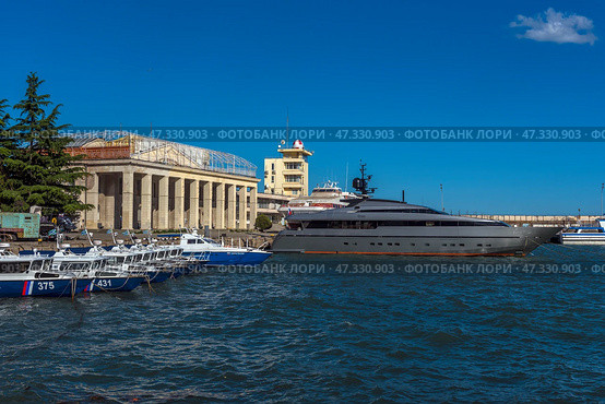 A fragment of the seaport in Yalta. In the center of the frame, an elegant and modern yacht called Orion is moored, painted dark gray with an orange line along the waterline. Nearby are boats and other vessels, and in the background are visible buildings of the port infrastructure, including the control tower with a characteristic dome. The sea is bright and rich blue, light ripples on the water indicate a fresh breeze. On the concrete wall behind is the inscription WELCOME, which emphasizes the tourist character of the place.The photo conveys the atmosphere of active marine life and the modern Yalta coast. (2021 год). Редакционное фото, фотограф Анна Кривицкая / Фотобанк Лори
