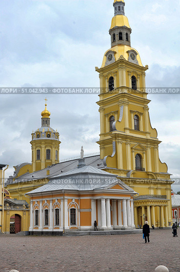 A general view of the Peter and Paul Cathedral in St. Petersburg, located on the territory of the Peter and Paul Fortress. In the foreground is a chapel with a colonnade and a triangular pediment, painted in warm orange and white tones. Behind it rises a majestic bell tower with a golden spire topped by an angel and a clock on the facade. On the right is the main dome of the cathedral with a golden cross and decorative elements in the Baroque style. The stone-paved square in front of the building gives the frame an atmosphere of old St. Petersburg. The weather is cloudy, which emphasizes the contrast between the gray sky and the bright yellow and gold facades. (2013 год). Редакционное фото, фотограф Анна Кривицкая / Фотобанк Лори