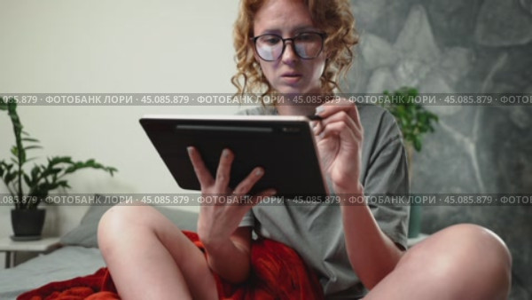 A girl with glasses is surfing the Internet at home in a cozy atmosphere and uses a tablet and a stylus. The glasses reflect the pages you scroll through on the Internet. Стоковое видео, видеограф Енваев Андрей / Фотобанк Лори