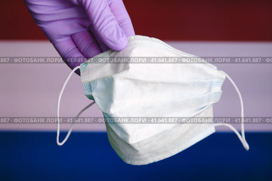 A hand glove holds a mask against the background of the Dutch flag. The fight against coronavirus in Netherlands. Стоковое фото, фотограф Андрей Журавлев / Фотобанк Лори