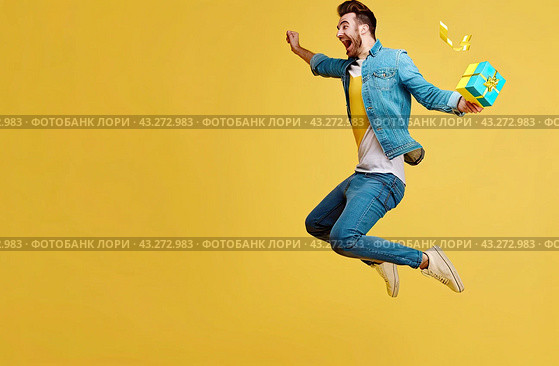 A happy young man with a gift box in his hand is jumping for joy on a yellow background. Copy space. Стоковое AI-изображение, AI-художник Ярославский Максим / Фотобанк Лори