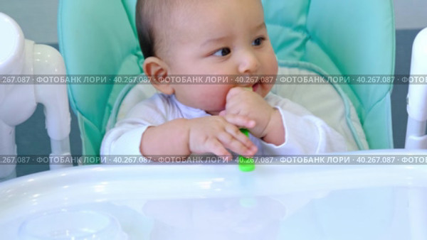 A hungry baby is gnawing on a plastic spoon at the table on a high chair. Teething, whims, itchy gums, introduction of complementary foods. Стоковое видео, видеограф Ольга Симонова / Фотобанк Лори