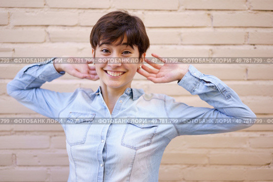 A joyful, vibrant young woman with short hair smiles and poses playfully against a light brick wall, radiating warmth, positivity, and confidence with her cheerful demeanor and carefree attitude. A Cheerful Young Woman with Short Hair Posing in Front of a Stylish Brick Wall Backdrop. Стоковое фото, агентство Ingram Publishing / Фотобанк Лори