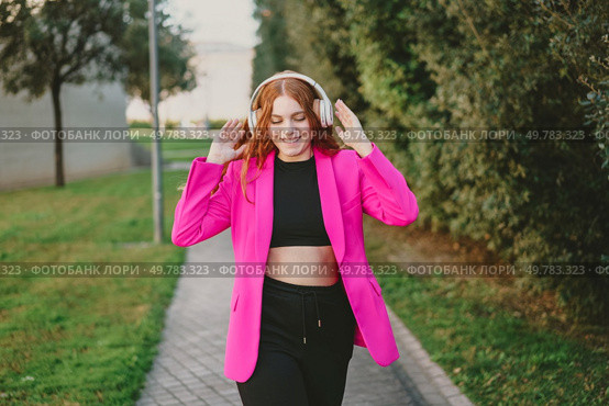 A joyful young woman enjoys her favorite music on a walk outdoors, wearing a vibrant pink blazer and stylish headphones, embodying modern youth and urban fashion in a lively atmosphere. A Happy Young Woman Enjoys Walking Outdoors While Wearing a Bright Pink Blazer and Headphones. Стоковое фото, агентство Ingram Publishing / Фотобанк Лори