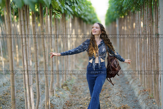 A joyful young woman with long hair strolls through a picturesque grove, savoring natures beauty and fresh air, radiating happiness and freedom, embracing the moment with delight. A Joyful Young Woman Happily Walking Among Tall Trees in Beautiful Nature Surroundings. Стоковое фото, агентство Ingram Publishing / Фотобанк Лори