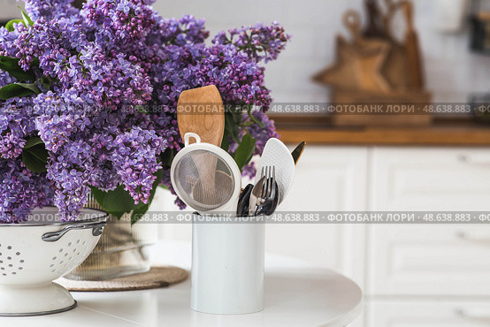 A large purple bouquet of lilacs and kitchen cutlery on the table. In the background is the interior of a modern white Scandinavian-style kitchen. The concept of home comfort and celebration. Стоковое фото, фотограф Photojuli / Фотобанк Лори