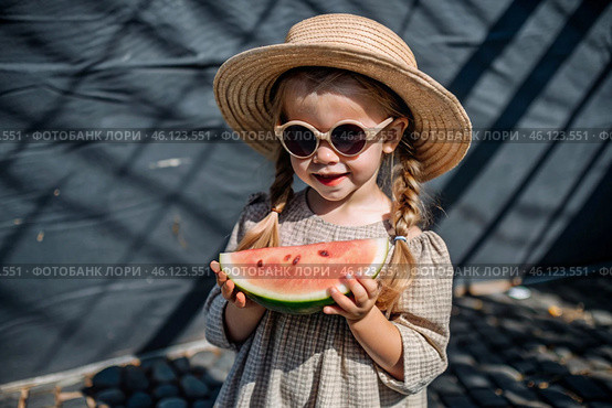 A little girl in sunglasses eats a watermelon in the hot summer. Funny happy child with a watermelon in his hands. Baby looks at the camera, healthy food. Стоковое AI-изображение, AI-художник Photojuli / Фотобанк Лори