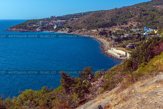 A magnificent view of the Crimean coast. From above, a panorama of the curved coastline, covered with sparse vegetation, rocks and junipers, opens up. In the distance, a small town or village with white houses spread out along the slope of a hill is visible. The Black Sea is deep blue, calm, with slight glare from the sun. On the horizon, closer to the center of the frame, an old sea platform or pier is noticeable. This place conveys an atmosphere of privacy, tranquility and southern warmth. Стоковое фото, фотограф Анна Кривицкая / Фотобанк Лори