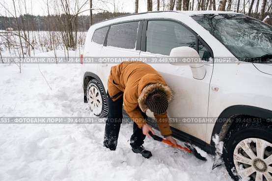 A man digs out a stalled car in the snow with a car shovel. Transport in winter got stuck in a snowdrift after a snowfall, sat on the bottom. First aid, tow truck, winter tires spikes and all-season. Стоковое фото, фотограф Ольга Симонова / Фотобанк Лори