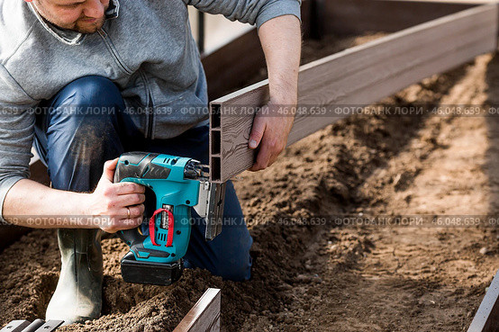 A man holds a battery-powered electric saw and builds new beds in a greenhouse made of wood-polymer composite. Macro photography. Side view. Preparing for the spring garden season. Стоковое фото, фотограф Photojuli / Фотобанк Лори