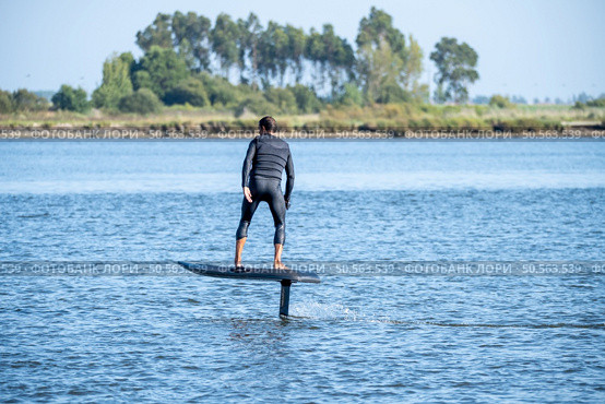 A man in a wetsuit rides a hydrofoil surfboard across calm waters, with trees lining the distant shore on a clear day. Стоковое фото, агентство Ingram Publishing / Фотобанк Лори