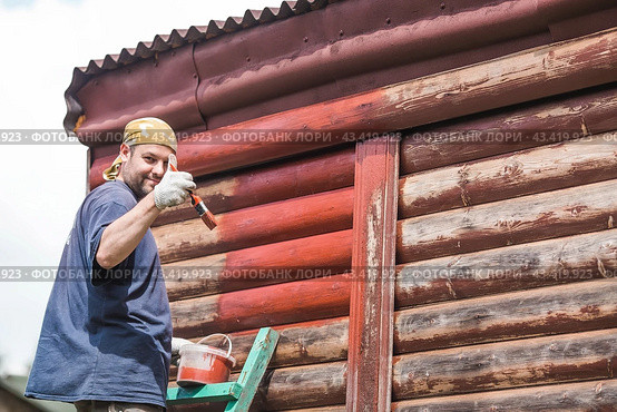 A man works with a brush and applies red paint to a wooden village house. A worker paints wooden planks in close-up. Half of the painted surface. Smearing with a brush. Стоковое фото, фотограф Photojuli / Фотобанк Лори
