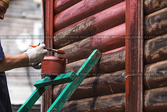 A man works with a brush and applies red paint to a wooden village house. A worker paints wooden planks in close-up. Half of the painted surface. Smearing with a brush. Стоковое фото, фотограф Photojuli / Фотобанк Лори